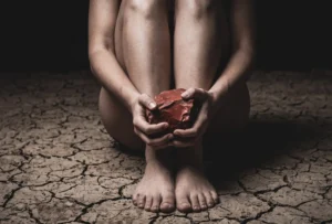 Tense posture on dry, cracked earth, holding raw Red Jasper, representing energy drain before grounding. |