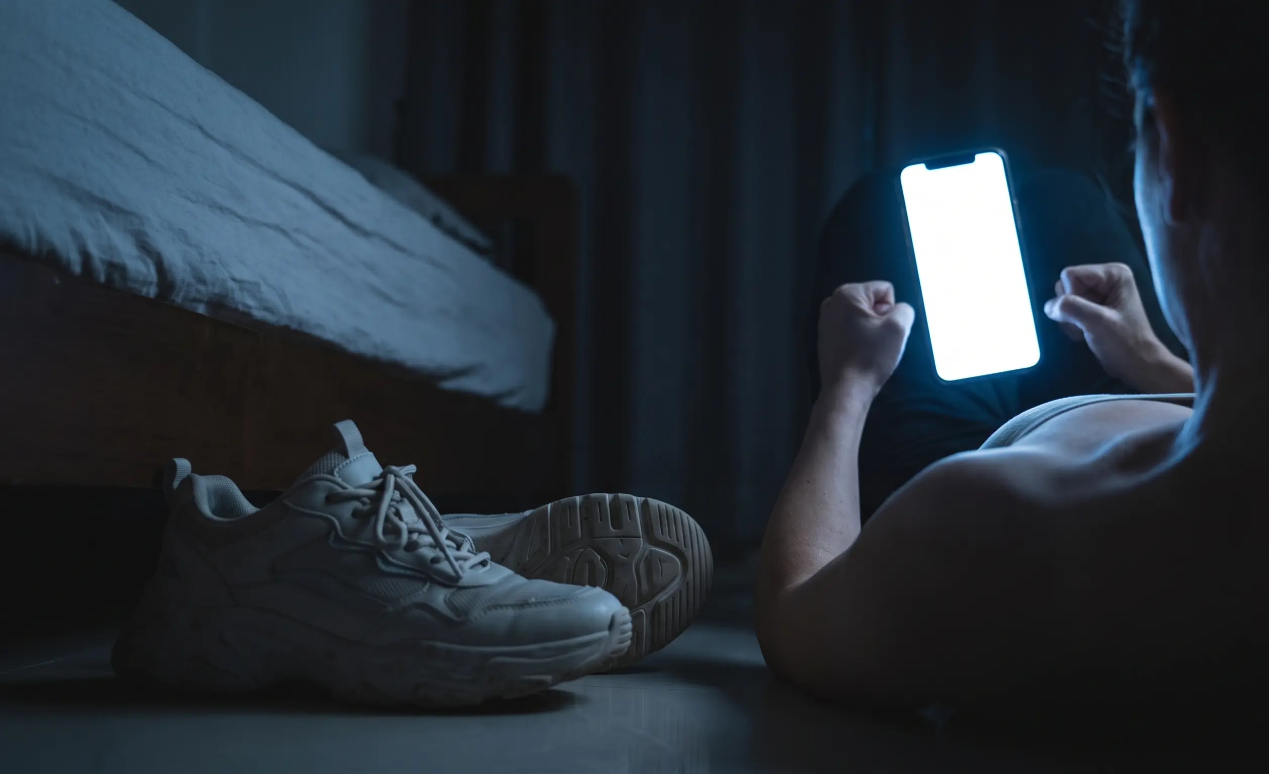 A close-up of thick synthetic shoes on a floor, next to a person viewed in the cold, blue-white static glow of a smartphone screen, visualizing urban energy overload.