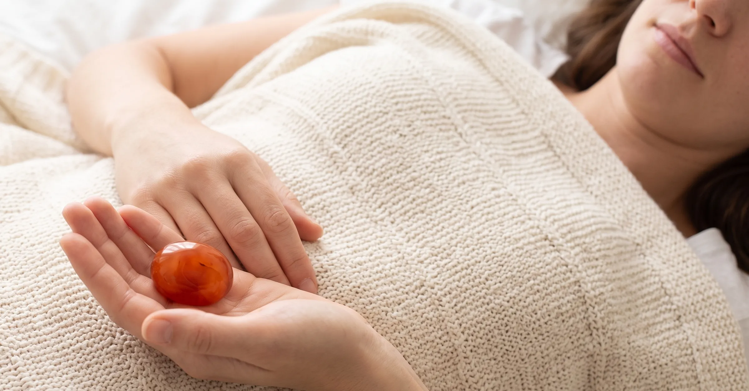 A person lying relaxed under a cream knitted blanket, holding a warm Carnelian stone gently over their lower abdomen.